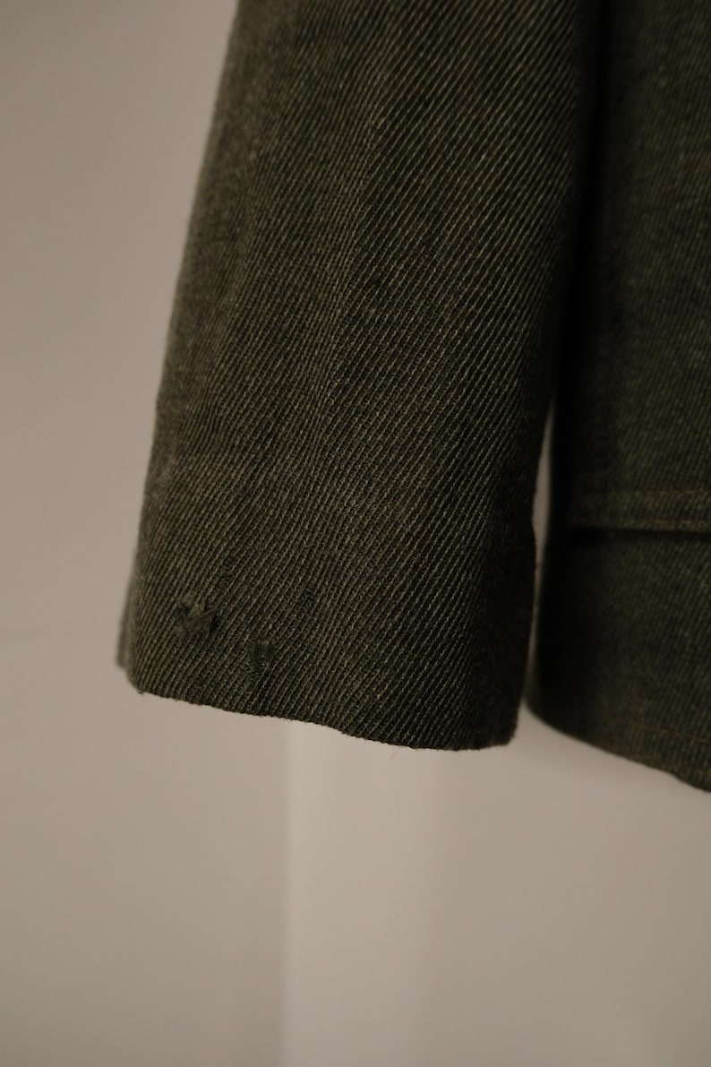 Puede incluir: Primer plano de una manga de chaqueta verde oscuro. La tela tiene un tejido diagonal texturizado. El pu&ntilde;o es visible, con unas peque&ntilde;as puntadas decorativas cerca del borde. La chaqueta parece estar hecha de un material duradero.