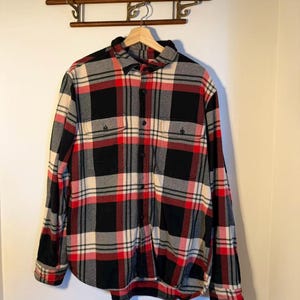 以下が含まれることがあります： 赤、黒、クリーム、グレーのチェック柄の長袖ボタンダウンフランネルシャツ。 シャツには胸ポケットが2つあり、木製のハンガーに掛けられています。 クラシックなスタイルです。