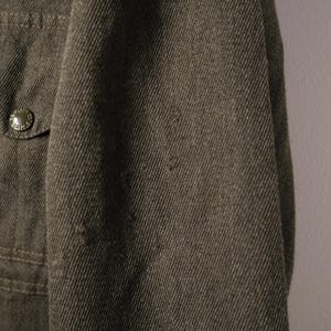 Puede incluir: Primer plano de una chaqueta verde oliva oscuro. La tela tiene un aspecto texturizado con l&iacute;neas diagonales visibles. Un peque&ntilde;o bolsillo con un bot&oacute;n es visible en el lado izquierdo. La chaqueta parece estar hecha de un material duradero.