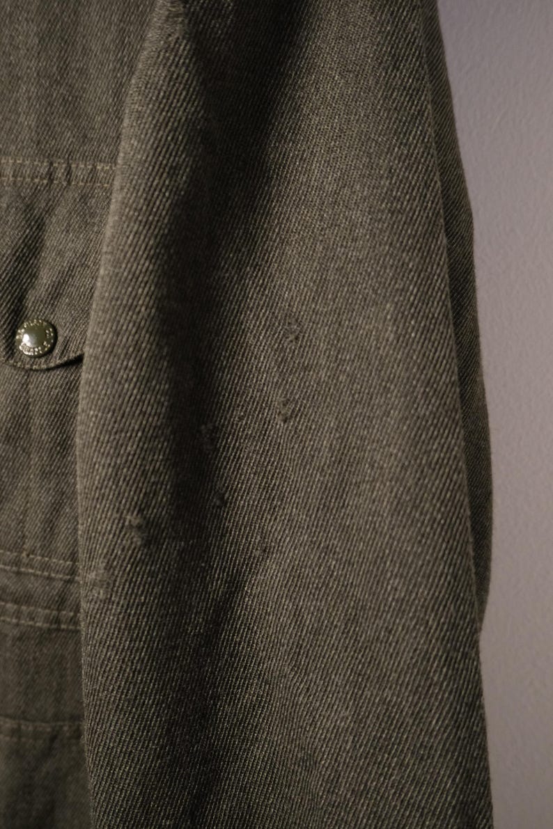 Puede incluir: Primer plano de una chaqueta verde oliva oscuro. La tela tiene un aspecto texturizado con l&iacute;neas diagonales visibles. Un peque&ntilde;o bolsillo con un bot&oacute;n es visible en el lado izquierdo. La chaqueta parece estar hecha de un material duradero.