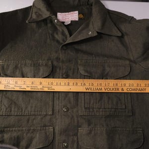 Puede incluir: Camisa verde oliva con botones, cuatro bolsillos delanteros y cuello. Una regla de madera est&aacute; colocada sobre la camisa, midiendo aproximadamente 66 cm. La camisa tiene una etiqueta que dice "FILSON GARMENT".