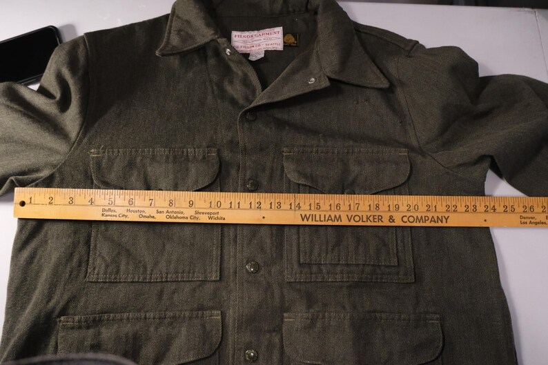 Puede incluir: Camisa verde oliva con botones, cuatro bolsillos delanteros y cuello. Una regla de madera est&aacute; colocada sobre la camisa, midiendo aproximadamente 66 cm. La camisa tiene una etiqueta que dice "FILSON GARMENT".