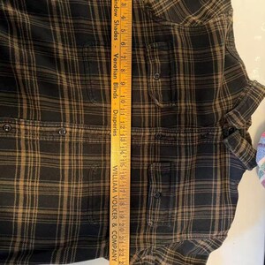 May include: A black and brown plaid flannel shirt with two chest pockets and button closures. A wooden ruler is placed vertically along the shirt, measuring approximately 26 inches in length. The shirt is folded.