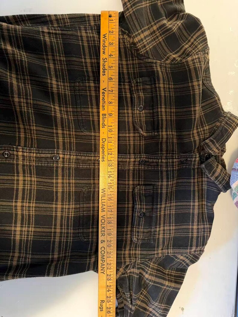 May include: A black and brown plaid flannel shirt with two chest pockets and button closures. A wooden ruler is placed vertically along the shirt, measuring approximately 26 inches in length. The shirt is folded.