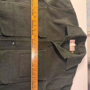 May include: A dark green wool jacket with four front pockets and a collared neckline. The jacket has button closures and a tag on the collar. The ruler shows the jacket is approximately 26 inches long.