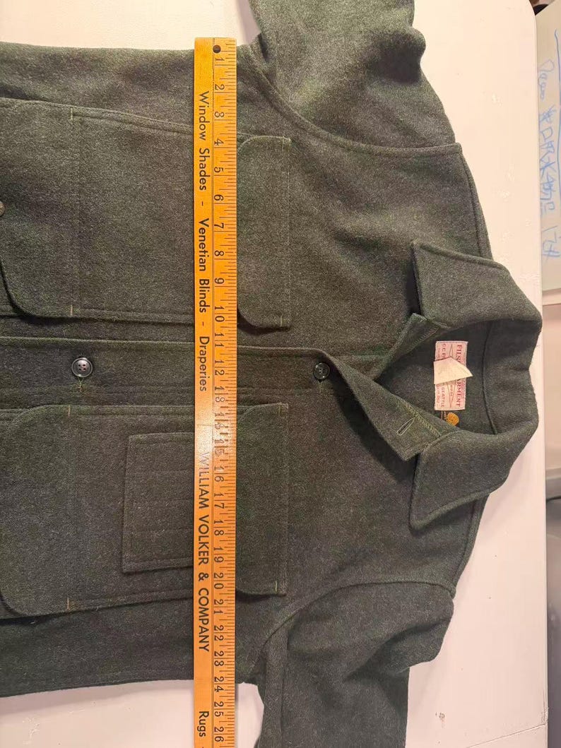 May include: A dark green wool jacket with four front pockets and a collared neckline. The jacket has button closures and a tag on the collar. The ruler shows the jacket is approximately 26 inches long.