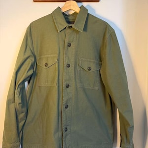 May include: Olive green button-up shirt with two chest pockets. The shirt has a collared neckline and long sleeves. The shirt is hanging on a wooden hanger.