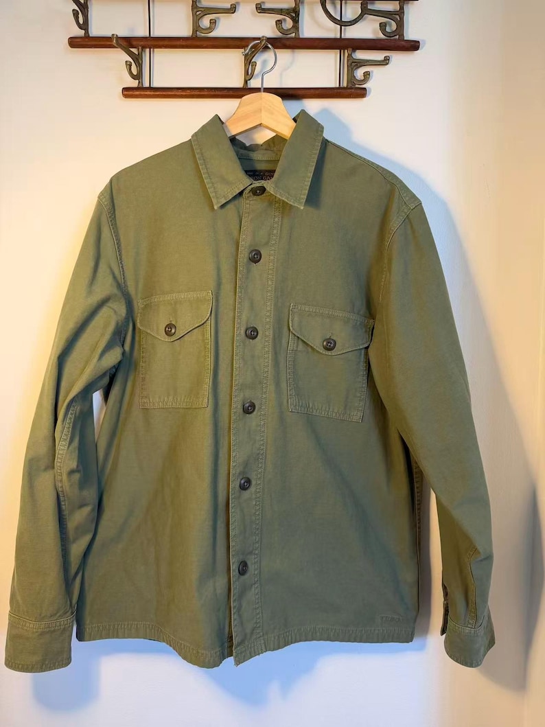 May include: Olive green button-up shirt with two chest pockets. The shirt has a collared neckline and long sleeves. The shirt is hanging on a wooden hanger.