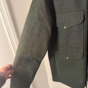 May include: Dark green wool jacket with two front pockets and button closures. The jacket has a classic design with a tailored fit. The sleeves are long and the fabric appears to be a heavy weight. The jacket is a solid color and suitable for cooler weather.