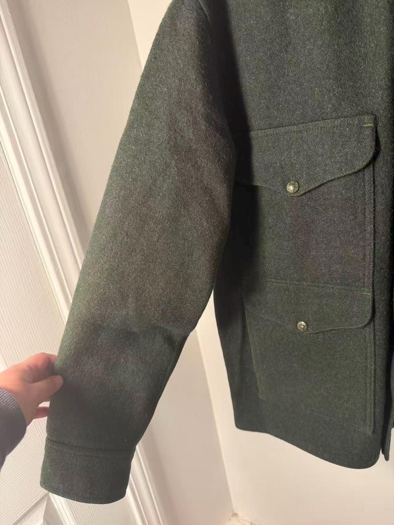 May include: Dark green wool jacket with two front pockets and button closures. The jacket has a classic design with a tailored fit. The sleeves are long and the fabric appears to be a heavy weight. The jacket is a solid color and suitable for cooler weather.