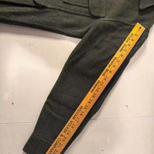 May include: Dark green wool jacket with a buttoned front, pockets, and a collar. A wooden ruler is placed along the sleeve, showing measurements in inches. The ruler has text that reads "Window Shades - Venetian Blinds - Draperies" and "WILLIAM VOLKER & COMPANY - Rugs."