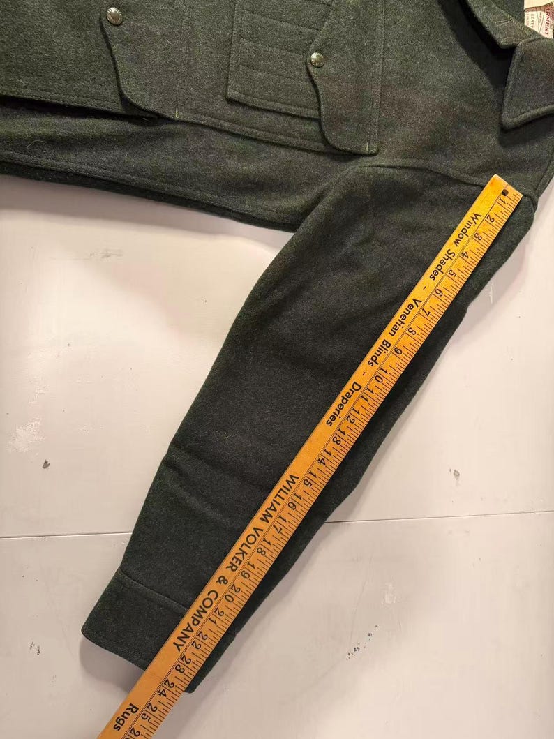 May include: Dark green wool jacket with a buttoned front, pockets, and a collar. A wooden ruler is placed along the sleeve, showing measurements in inches. The ruler has text that reads "Window Shades - Venetian Blinds - Draperies" and "WILLIAM VOLKER & COMPANY - Rugs."