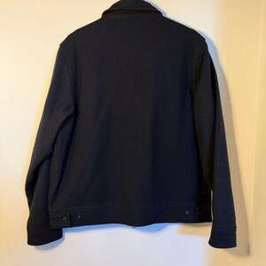 Puede incluir: Una chaqueta azul marino con cuello y dos bolsillos abotonados en la cintura. La chaqueta est&aacute; colgada en un perchero de madera. La chaqueta parece estar hecha de un material grueso y c&aacute;lido.