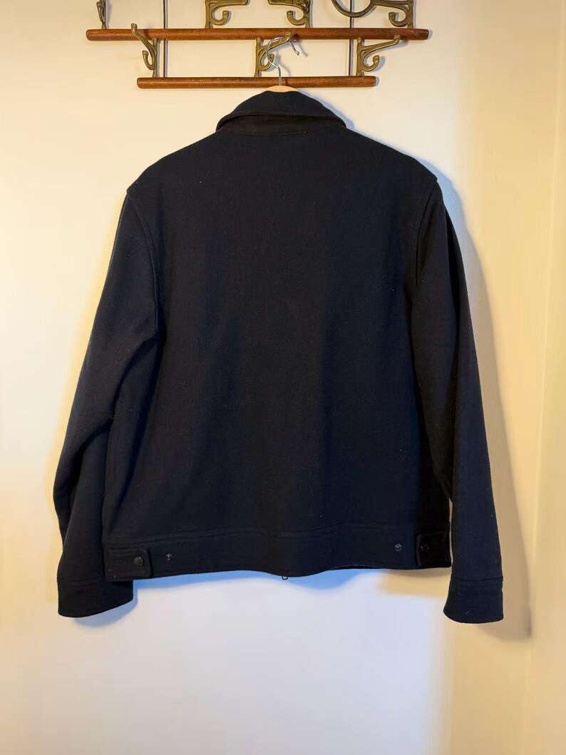 Puede incluir: Una chaqueta azul marino con cuello y dos bolsillos abotonados en la cintura. La chaqueta est&aacute; colgada en un perchero de madera. La chaqueta parece estar hecha de un material grueso y c&aacute;lido.