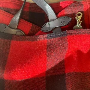 Op de afbeelding: Close-up van een rood-zwarte geruite stof met lederen details. De afbeelding toont een zak met een goudkleurige sluiting en leren riemen. De stof heeft een getextureerd uiterlijk.