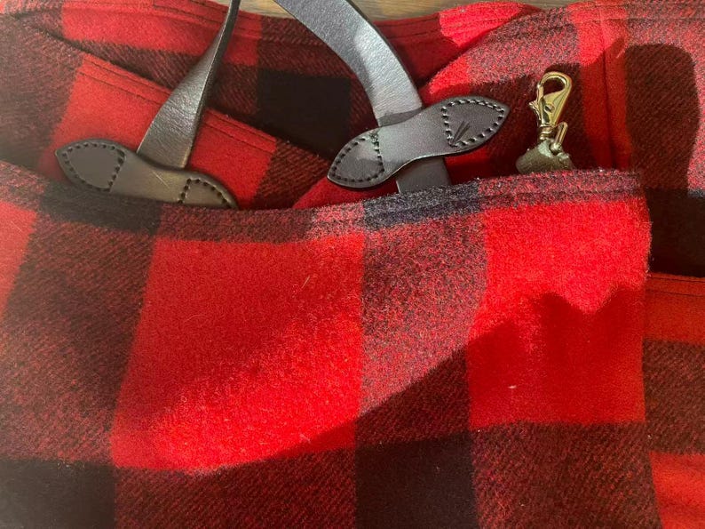 Op de afbeelding: Close-up van een rood-zwarte geruite stof met lederen details. De afbeelding toont een zak met een goudkleurige sluiting en leren riemen. De stof heeft een getextureerd uiterlijk.