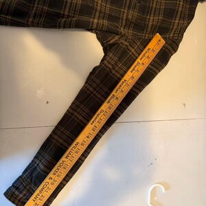 May include: A dark green and brown plaid flannel shirt with long sleeves. A wooden ruler is placed on the sleeve, measuring up to 27 inches. The shirt is lying on a white surface, with a white hanger in the bottom right corner.