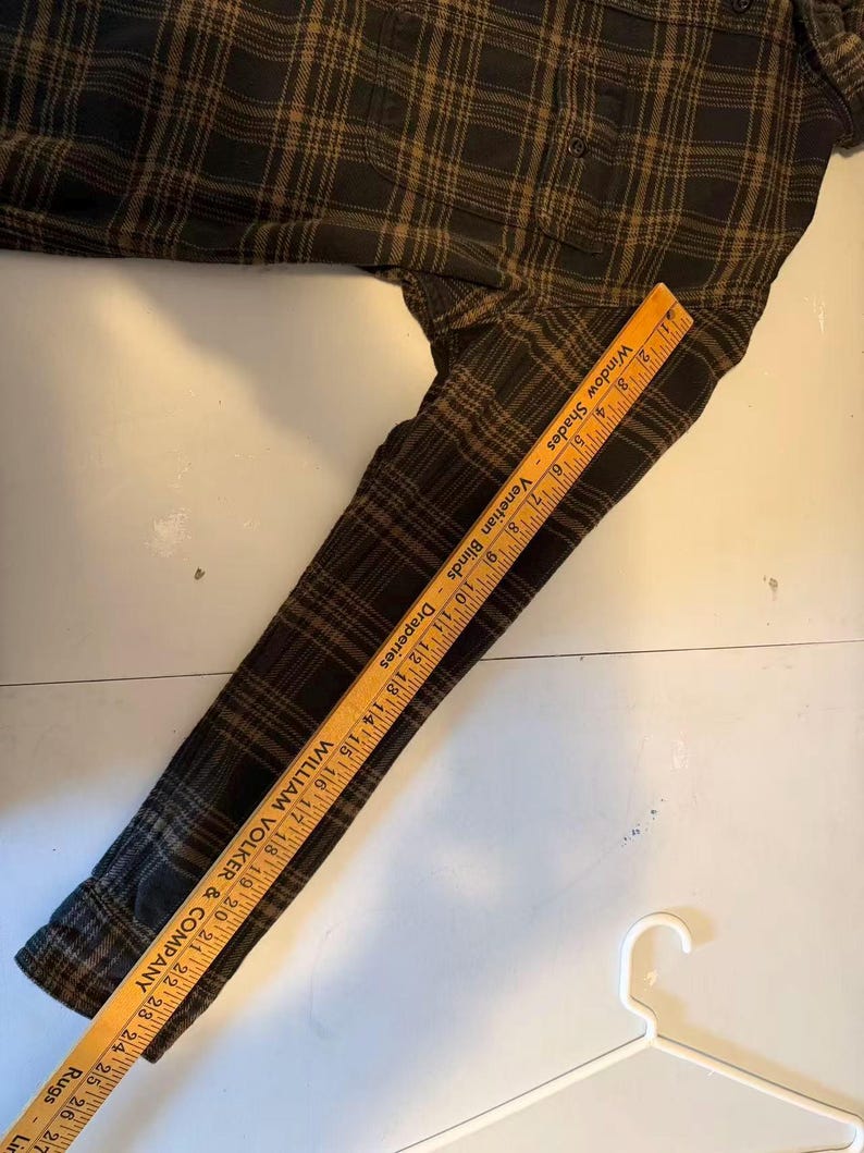 May include: A dark green and brown plaid flannel shirt with long sleeves. A wooden ruler is placed on the sleeve, measuring up to 27 inches. The shirt is lying on a white surface, with a white hanger in the bottom right corner.