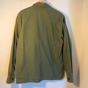 May include: Olive green long-sleeve shirt hanging on a wooden hanger. The shirt has a button-down front, a collar, and buttoned cuffs. The back of the shirt is visible, showing the seams and the texture of the fabric.