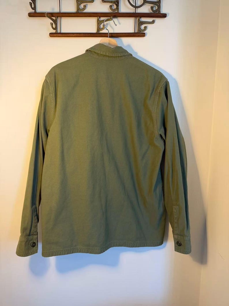 May include: Olive green long-sleeve shirt hanging on a wooden hanger. The shirt has a button-down front, a collar, and buttoned cuffs. The back of the shirt is visible, showing the seams and the texture of the fabric.