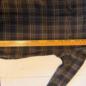 May include: A dark plaid button-down shirt with a ruler measuring its length. The shirt features a classic plaid pattern in shades of black and brown. The ruler shows a measurement of approximately 10 inches.
