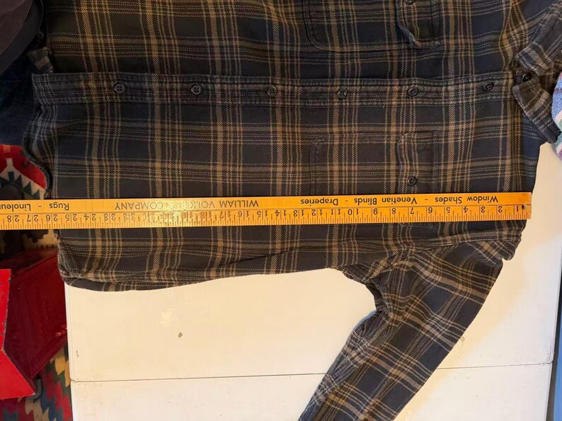 May include: A dark plaid button-down shirt with a ruler measuring its length. The shirt features a classic plaid pattern in shades of black and brown. The ruler shows a measurement of approximately 10 inches.