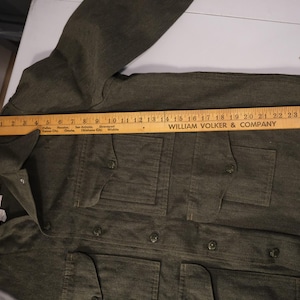 Puede incluir: Una chaqueta verde oliva oscuro con cuatro bolsillos delanteros y cierres de botones. Una regla de madera est&aacute; colocada sobre la chaqueta, midiendo aproximadamente 71 cm de largo. La chaqueta tiene cuello y mangas largas.