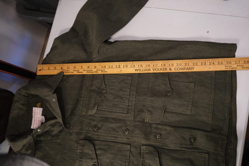 Puede incluir: Una chaqueta verde oliva oscuro con cuatro bolsillos delanteros y cierres de botones. Una regla de madera est&aacute; colocada sobre la chaqueta, midiendo aproximadamente 71 cm de largo. La chaqueta tiene cuello y mangas largas.