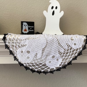 May include: A white crocheted Halloween doily with a black scalloped edge. The doily features ghost-shaped cutouts. A small black sign with the text "Beware of Ghosts" and a wooden ghost decoration are also visible.