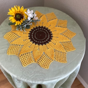 May include: A crocheted sunflower table runner in yellow and brown. The runner is on a light green tablecloth with a floral pattern.