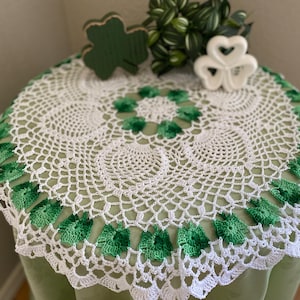 May include: White crochet table runner with a green shamrock design around the edge. The runner is on a green table with a wooden shamrock and a white ceramic shamrock on the table.