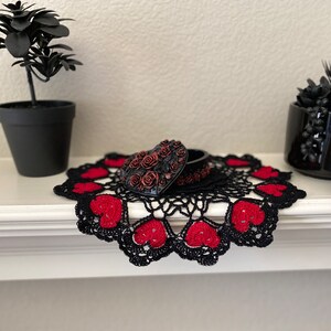 May include: A black heart-shaped trinket box with red rose details sits on a black crocheted doily with red hearts. The doily is on a white surface.