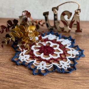 May include: A handmade, crocheted coaster in red, white, and blue, with a scalloped edge. The coaster is placed on a wooden surface, with a decorative "USA" sign in the background. A small floral arrangement is also present.