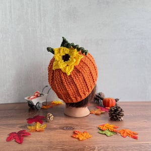 May include: A crocheted pumpkin-shaped hat in a vibrant orange color, featuring a textured ribbed pattern. The top of the hat is adorned with a large, bright yellow crocheted sunflower with a dark brown center and green crocheted leaves.