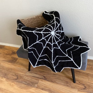May include: A black and white crocheted blanket with a spiderweb design. The blanket is draped over a small, gray upholstered bench with dark wooden legs. The spiderweb pattern is white against a black background.