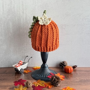 May include: An orange crochet pumpkin hat with green and cream-colored leaf accents. The hat is displayed on a black stand, surrounded by fall-themed decorations like leaves and a small pumpkin. The hat is a seasonal accessory.