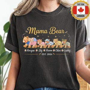 May include: Dark grey t-shirt featuring the text "Mama Bear" in gold script above a row of cartoon bears. Each bear wears different outfits and has a name below. The names are Morgan, Lily, Mason, Chloe, and Gabby. The text "EST. 2026" is below the names.
