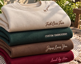 May include: A stack of four crewneck sweatshirts in beige, dark green, brown, and maroon. The beige sweatshirt features the text by grace and Faith Over Fear. The dark green one says CUSTOM EMBROIDERY, the brown says Jesus Loves You, and the maroon says loved by Him.