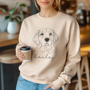May include: A beige sweatshirt with a line drawing of a golden retriever dog and the name "Bailey" printed on the front. The left sleeve has the text "DOG MOM" and a small heart graphic.