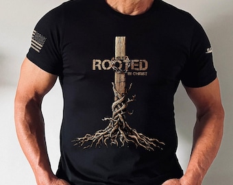 Puede incluir: Una camiseta negra con un gráfico de una cruz de madera con raíces y el texto "Rooted in Christ".