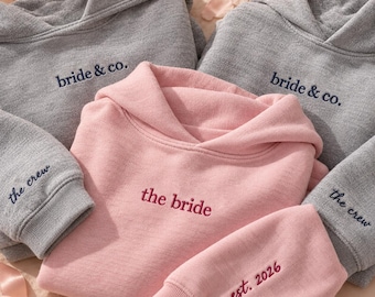 May include: Two heather gray hoodies with dark blue embroidery reading bride & co. and the crew on the sleeves, alongside a light pink hoodie with dark pink embroidery reading the bride and est. 2026 on the sleeve, all featuring hoods.
