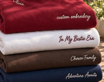 May include: A stack of four crewneck sweatshirts in maroon, white, brown, and navy blue. The maroon sweatshirt features the text Olivia and custom embroidery. The white sweatshirt reads In My Bestie Era. The brown reads Chosen Family, and the navy blue reads Adventure Awaits.