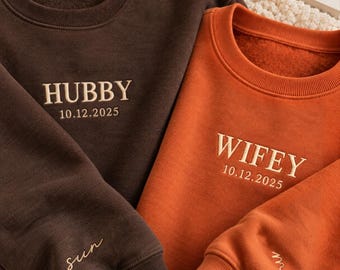 May include: A dark brown sweatshirt embroidered with HUBBY and 10.12.2025 on the chest and sun on the sleeve, paired with an orange sweatshirt embroidered with WIFEY and 10.12.2025 on the chest and moon on the sleeve, both with crew necks.