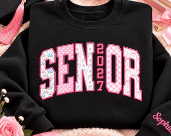 May include: A black crewneck sweatshirt featuring the word SENIOR with the year 2027 in the center. The letters are filled with various pink and blue floral and gingham patterns, outlined in pink. The name Sophia is embroidered on the sleeve.