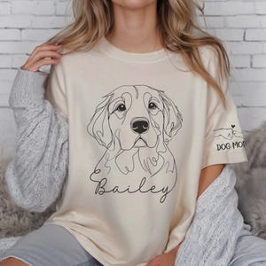 May include: A beige t-shirt with a line drawing of a golden retriever dog and the name "Bailey" printed on the front. The shirt has a rolled-up sleeve with the text "Dog Mom" printed on it.