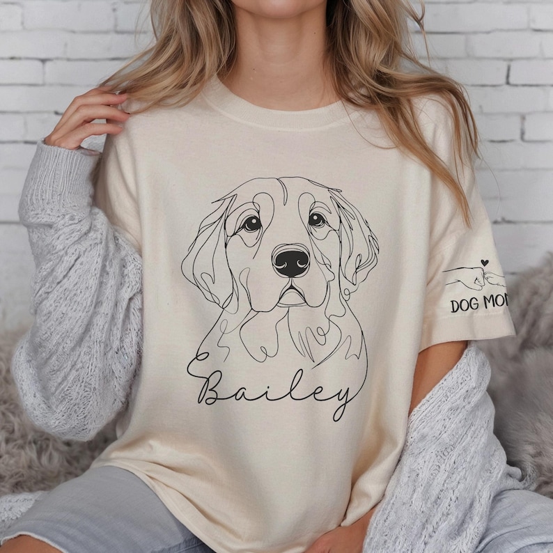 May include: A beige t-shirt with a line drawing of a golden retriever dog and the name "Bailey" printed on the front. The shirt has a rolled-up sleeve with the text "Dog Mom" printed on it.