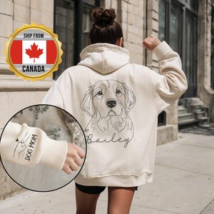 Peut inclure: Sweat à capuche crème avec un dessin au trait de chien et le nom "Bailey". Le poignet gauche affiche "DOG MOM" avec une empreinte de patte et un cœur. Le sweat à capuche a un col montant.