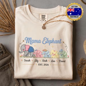 May include: Cream-coloured t-shirt with the text "Mama Elephant" in blue script, featuring embroidered elephants. Below are names and "EST. 2026". A gold necklace is draped on the shirt. The shirt is from Australia.