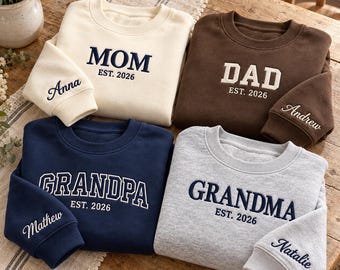 May include: Four crewneck sweatshirts in cream, brown, navy blue, and heather gray. Each features embroidered text: MOM EST. 2026, DAD EST. 2026, GRANDPA EST. 2026, and GRANDMA EST. 2026. The sleeves are embroidered with the names Anna, Andrew, Mathew, and Natalie.