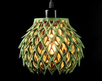 May include: A sage green pendant light fixture featuring a complex, three-dimensional lattice structure with a repeating diamond-shaped pattern. The shade is suspended from a black cord and emits a warm, glowing light from the center bulb.